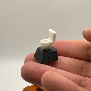 Puede incluir: Un inodoro blanco en miniatura con una base negra, sentado en la mano de una persona.