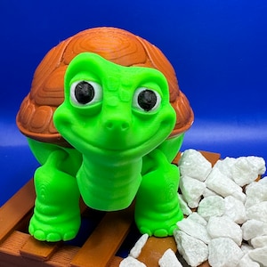 May include: A green turtle figurine with a brown shell, sitting on a wooden platform with white rocks. The turtle has large, black eyes and a friendly expression.