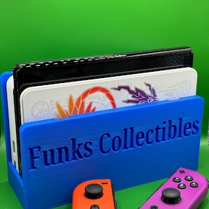 May include: A blue 3D printed stand with the text "Funks Collectibles" on the front. The stand holds a Nintendo Switch console and two controllers. One controller is red and the other is purple.