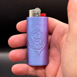 May include: A purple and pink lighter with a textured finish and an embossed design of two fish. The lighter has a silver top and a red button.