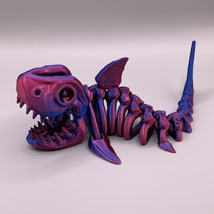 May include: A 3D printed shark skeleton figurine with a purple and blue iridescent finish. The figurine is posed with its mouth open and its tail raised.