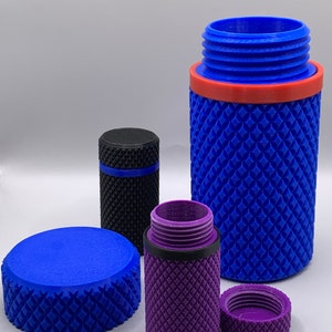 Op de afbeelding: Drie 3D-geprinte containers, een blauwe, een paarse en een zwarte. De blauwe container is open en heeft een rode ring rond de bovenkant. De paarse container is open en heeft een zwarte ring rond de bovenkant. De zwarte container is gesloten en heeft een blauwe ring rond de bovenkant.