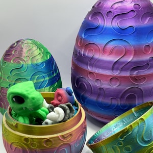 May include: Three colorful, patterned, plastic Easter eggs. The smallest egg is open and contains a green and purple toy. The largest egg is closed and has a purple, blue, and pink color scheme. The middle egg is open and has a yellow, pink, and blue color scheme.