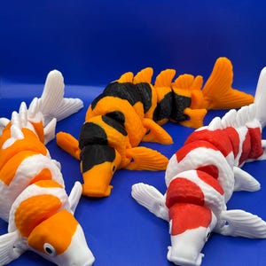 May include: Three colorful koi fish figurines in orange, white, black, and red are arranged on a blue surface. The fish have detailed fins and scales, showcasing a vibrant and decorative design. The koi are a popular symbol of good fortune.
