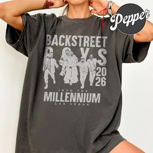 Pode incluir: Uma t-shirt de manga curta em cinzento-carvão com um gráfico branco desgastado dos Backstreet Boys. A impressão inclui o texto Backstreet Boys, Into The Millennium, Las Vegas e o ano 2026, juntamente com silhuetas dos membros da banda.