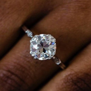 May include: A close-up of a hand wearing a silver ring with a large, round, clear diamond. The ring has a delicate band with small diamonds on either side of the center stone.