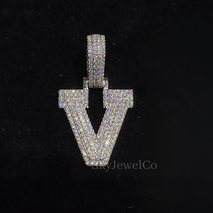 May include: A silver-colored pendant in the shape of the letter "V", encrusted with small, sparkling stones. The pendant hangs from a matching bail, and is set against a black background. The brand name "Sky JewelCo" is visible.
