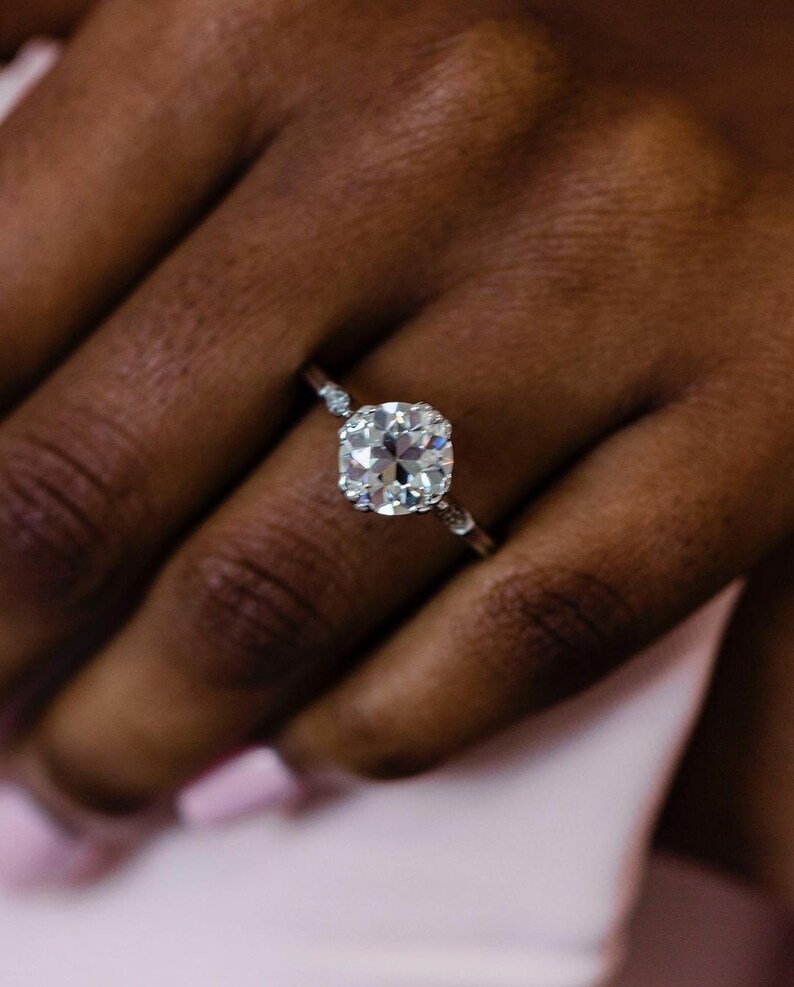 May include: A close-up of a hand wearing a silver ring with a large, round, clear diamond. The ring has a delicate band with small diamonds on either side of the center stone.
