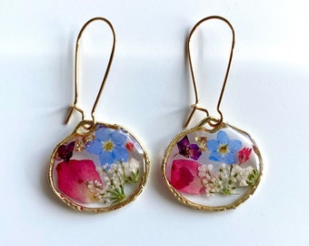 Pode incluir: Um par de brincos de ouro com um pingente oval de resina transparente contendo flores prensadas. As flores são rosa, azul e branco.