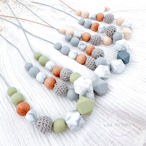 May include: A collection of silicone teething necklaces in various colors and textures, including green, gray, white, and brown. The necklaces feature round, square, and textured beads, as well as crocheted beads. The necklaces are displayed on a white surface. "LOVE MY BABY" is written on a pink circle in the bottom right corner of the image.