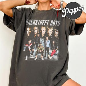 Pode incluir: Uma t-shirt de manga curta cinzento-carvão com uma impressão gráfica de estilo vintage dos Backstreet Boys. O design inclui uma colagem dos membros da banda em várias poses com o texto BACKSTREET BOYS em letras maiúsculas brancas.
