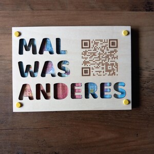 Könnte beinhalten: Ein Holzschild mit dem deutschen Satz "MAL WAS ANDERES" ausgeschnitten, der farbige Euro-Banknoten zeigt. Auf der rechten Seite ist ein QR-Code eingeätzt, mit gelben Akzenten in den Ecken. Das Schild liegt auf einer Holzoberfläche.