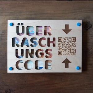 May include: A light-coloured wooden sign with the German words "ÜBER RASCH UNGS CODE" cut out, revealing currency. A QR code and arrows are also present. The sign is held together with four blue tacks.