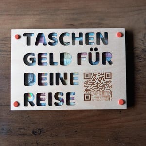 Könnte beinhalten: Ein Holzschild mit dem ausgeschnittenen deutschen Text "TASCHEN GELD FÜR DEINE REISE", der farbenfrohe Währungen zeigt. Das Schild hat einen QR-Code und orangefarbene runde Stifte.