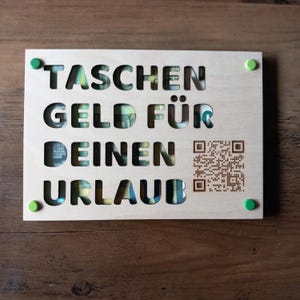 Può includere: Un cartello di legno con il testo tedesco "TASCHENGELD FÜR DEINEN URLAUB" ritagliato, che rivela denaro all'interno. Il cartello ha dettagli verdi e gialli e un codice QR.