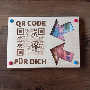 Könnte beinhalten: Helle Holzplakette mit QR-Code und den Worten "QR CODE" und "FÜR DICH" eingraviert. Zwei pfeilförmige Ausschnitte zeigen farbenfrohe Währung. Rote und blaue Reißzwecken befinden sich in jeder Ecke.
