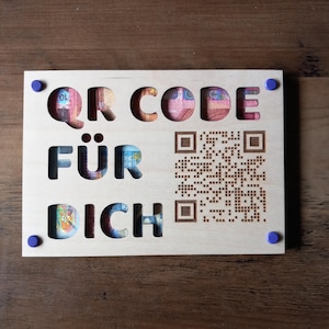 Könnte beinhalten: Eine Holzplakette mit den ausgeschnittenen Worten "QR CODE FÜR DICH", die farbenfrohe Banknoten zeigt. Auf der rechten Seite befindet sich ein QR-Code. Die Plakette ist mit blauen Befestigungen gesichert. Der Hintergrund ist eine dunkle Holzoberfläche.