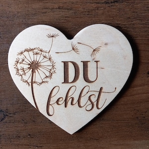 Könnte beinhalten: Ein herzförmiges Schild aus Holz mit dem Text "DU fehlst" und einem Löwenzahnmuster.