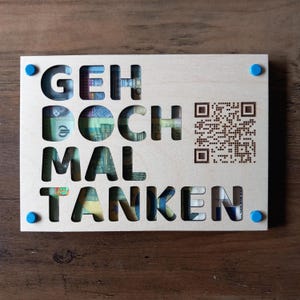 Puede incluir: Un letrero de madera clara con la frase alemana "GEH DOCH MAL TANKEN" recortada, que revela moneda. El letrero tiene un código QR y detalles en azul.