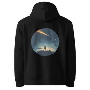 May include: Black hooded sweatshirt featuring a circular graphic on the back. The graphic depicts a silhouette of a person standing on a hill, gazing at a starry night sky with a shooting star. The design includes shades of blue, yellow, and white.