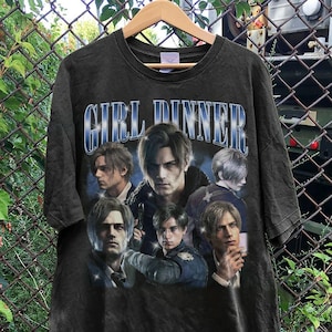 May include: A black short-sleeve t-shirt featuring a graphic print of multiple images of a male character with blonde hair. The text "GIRL DINNER" is printed in a large, stylized blue font with a white outline at the top of the graphic design.
