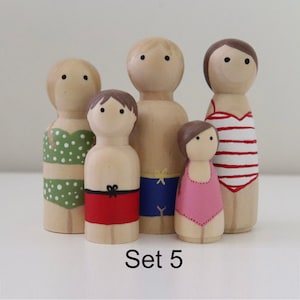 Pode incluir: Conjunto de cinco bonecas de madeira com pinos vestidas com roupas de banho. As bonecas são pintadas em várias cores, incluindo verde, vermelho, azul, rosa e branco. As bonecas usam todos diferentes estilos de roupas de banho, incluindo um maiô, um biquíni e sungas. As bonecas estão em pé sobre uma superfície branca. O texto "Set 5" está impresso abaixo das bonecas.