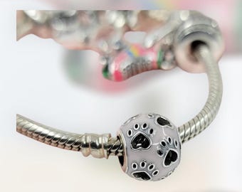 Peut inclure: Une perle de breloque rose clair avec des empreintes de pattes noires et des accents d'émail blanc. La breloque est sur un bracelet en argent. La perle est ronde et a une finition brillante. Accessoire sur le thème des animaux de compagnie.