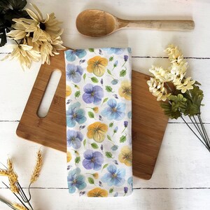 Puede incluir: Un paño de cocina blanco con un estampado floral de pensamientos azules, amarillos y morados. El paño está sobre una tabla de cortar de madera, con una cuchara de madera y flores en el fondo. El paño mide aproximadamente 50 cm de largo.