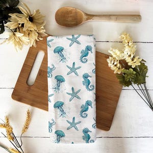 Puede incluir: Toalla de cocina blanca con un dise&ntilde;o de acuarela azul de medusas, caballitos de mar y estrellas de mar. La toalla est&aacute; sobre una tabla de cortar de madera con una cuchara de madera y flores.