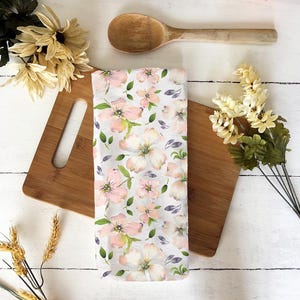 Puede incluir: Un paño de cocina blanco con un estampado floral de flores rosas y blancas y hojas verdes. El paño está sobre una tabla de cortar de madera, con una cuchara de madera y flores en el fondo.