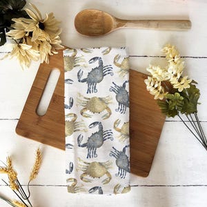 Puede incluir: Un paño de cocina blanco con un patrón repetido de cangrejos de acuarela en tonos azules y beige. El paño se muestra en una tabla de cortar de madera con una cuchara de madera y flores decorativas.