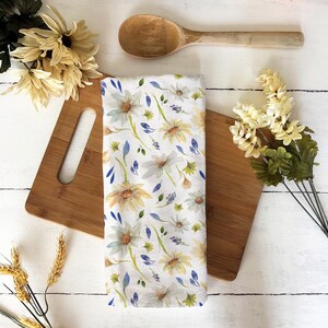 Puede incluir: Un paño de cocina blanco con un estampado floral de margaritas amarillas y blancas, hojas azules y tallos verdes. El paño está colocado sobre una tabla de cortar de madera, con una cuchara de madera y flores.