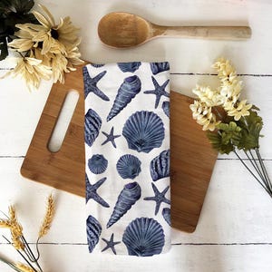 Puede incluir: Un paño de cocina blanco con un estampado de conchas marinas y estrellas de mar azules. El paño está sobre una tabla de cortar de madera, con una cuchara de madera y flores en el fondo. El paño es de un material suave y absorbente.