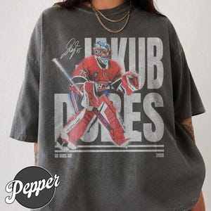 Op de afbeelding: Een antracietgrijs T-shirt met korte mouwen en een afbeelding van een ijshockeykeeper in rood en blauw tenue. De tekst luidt JAKUB DUBES, GO HABS GO en 2026. Boven de keeper staat een handtekening. Het shirt heeft een vintage, verweerde look.