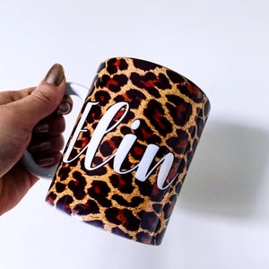 May include: A white ceramic mug with a leopard print design and the name "Eilin" written in white cursive lettering.
