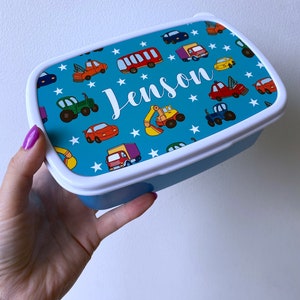 May include: A blue lunchbox with a white lid and a colorful design of cars, trucks, and stars. The name "Jenson" is written in white on the front of the lunchbox.