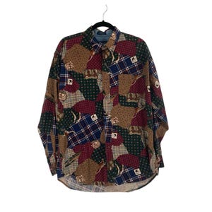 May include: A long-sleeved button-down shirt with a patchwork design. The shirt features a mix of patterns, including plaid, polka dots, and paisley, in shades of burgundy, navy, green, and tan. The shirt is hanging on a black hanger.