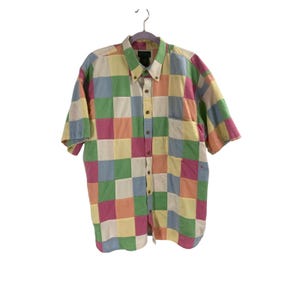 May include: A short-sleeved button-down shirt in a patchwork design. The shirt features a variety of pastel colors, including pink, yellow, green, and blue squares. It has a button-down collar and a single chest pocket.