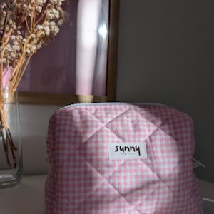 Könnte beinhalten: Rosa-weiß karierte, gesteppte Kosmetiktasche mit Reißverschluss. Die Tasche hat ein Etikett mit der Aufschrift "sunny". Die Tasche liegt auf einer weißen Oberfläche.