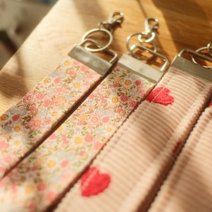 Könnte beinhalten: Drei Schlüsselanhänger aus Stoff mit silberfarbenen Metallbeschlägen und Karabinerhaken. Einer zeigt ein mehrfarbiges Blumenmuster auf hellem Grund, während die anderen beiden aus hellrosa Cordstoff mit kleinen roten Stickherzen bestehen.