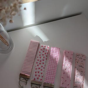 Könnte beinhalten: Fünf rosa Schlüsselanhänger aus Stoff mit silberfarbener Hardware. Die Schlüsselanhänger weisen verschiedene Muster auf, darunter Streifen, Blumenmuster, Vichy-Karo und Fruchtdesigns. Die Schlüsselanhänger werden auf einer weißen Oberfläche präsentiert.
