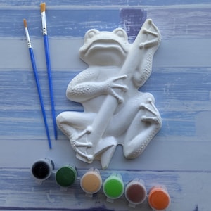 May include: A white plaster frog figurine holding a stick, accompanied by two blue-handled paintbrushes and a strip of six small paint pots containing black, dark green, tan, light green, brown, and orange paint colours for crafting.