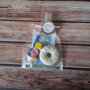 May include: A clear plastic bag containing a paint-your-own donut kit. The kit includes a white ceramic donut, four small pots of paint, a paintbrush, and a card with a rainbow design.