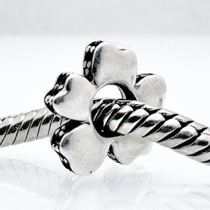 Puede incluir: Un charm de plata con un diseño de trébol de cuatro hojas. El charm está ensartado en una pulsera de cadena de serpiente plateada. El trébol tiene una superficie texturizada con pequeños acentos oscuros. La pulsera tiene un patrón trenzado.