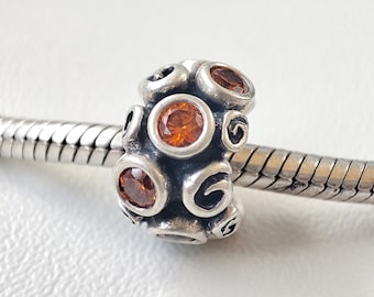 May include: A silver charm with multiple orange-colored gemstones. The charm is cylindrical with a series of circular and oval-shaped orange stones. The charm is on a silver chain.