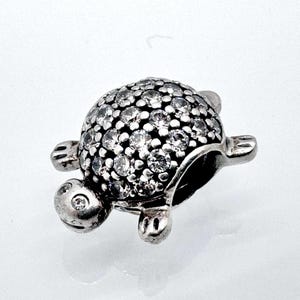 Può includere: Un ciondolo a forma di tartaruga argentata con un guscio ricoperto di piccole pietre chiare e scintillanti. La tartaruga ha piccole zampe, una testa con due occhi e un corpo liscio e arrotondato. Il ciondolo è probabilmente per un braccialetto o una collana.