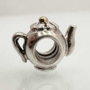 Peut inclure: Charme en forme de théière en argent avec un détail de fleur dorée. Le charme a un trou au centre pour être enfilé sur un bracelet.