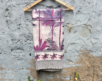 May include: A white cotton fabric with a pink and purple palm tree print hanging on a wooden hanger against a gray wall.