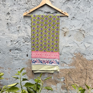 Może przedstawiać: Zielony bawełniany materiał z różowym i niebieskim nadrukiem kwiatowym wisi na drewnianym wieszaku na tle szorstkiej szarej ściany. Materiał ma białą obwódkę z paskowanym wzorem.