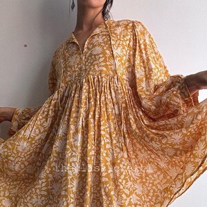 May include: Mustard yellow dress with a white floral pattern. The dress has long sleeves, a collared neckline, and a flowing skirt. The hemline falls above the knees. The dress is paired with brown cowboy boots.